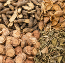 Four distinct piles of natural whole botanical ingredients arranged in a neat quadrant pattern, featuring dark brown roots, sliced tan roots, dried orange-brown seeds, and greenish-brown dried leaves.