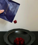 A hand holds a dark blue pouch labeled 'LUMIDROPS' above a black bowl containing several red dome-shape gummy, with one candy floating mid-air.