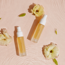 Two frosted glass spray bottles filled with golden-yellow liquid, arranged on a soft pink surface with ripples and pale flowers around them.