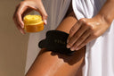 A person applying yellow cream from a  YINA Recovery Body Treatment jar onto their leg using a black YINA Bian Stone gua sha massage tool, with a white robe draped around them.